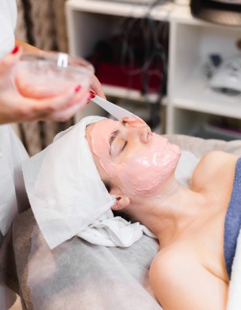 Cosmetologist applies alginate mask with spatula on face of woman. Facial skin and anti-aging treatment. Cosmetology and professional facial skin care.